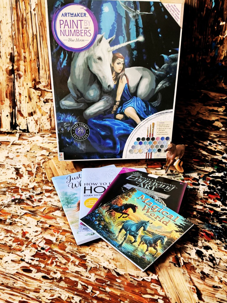 A paint by numbers kit featuring a woman in a blue dress beside a white unicorn, alongside instructional art books about horses and drawing.